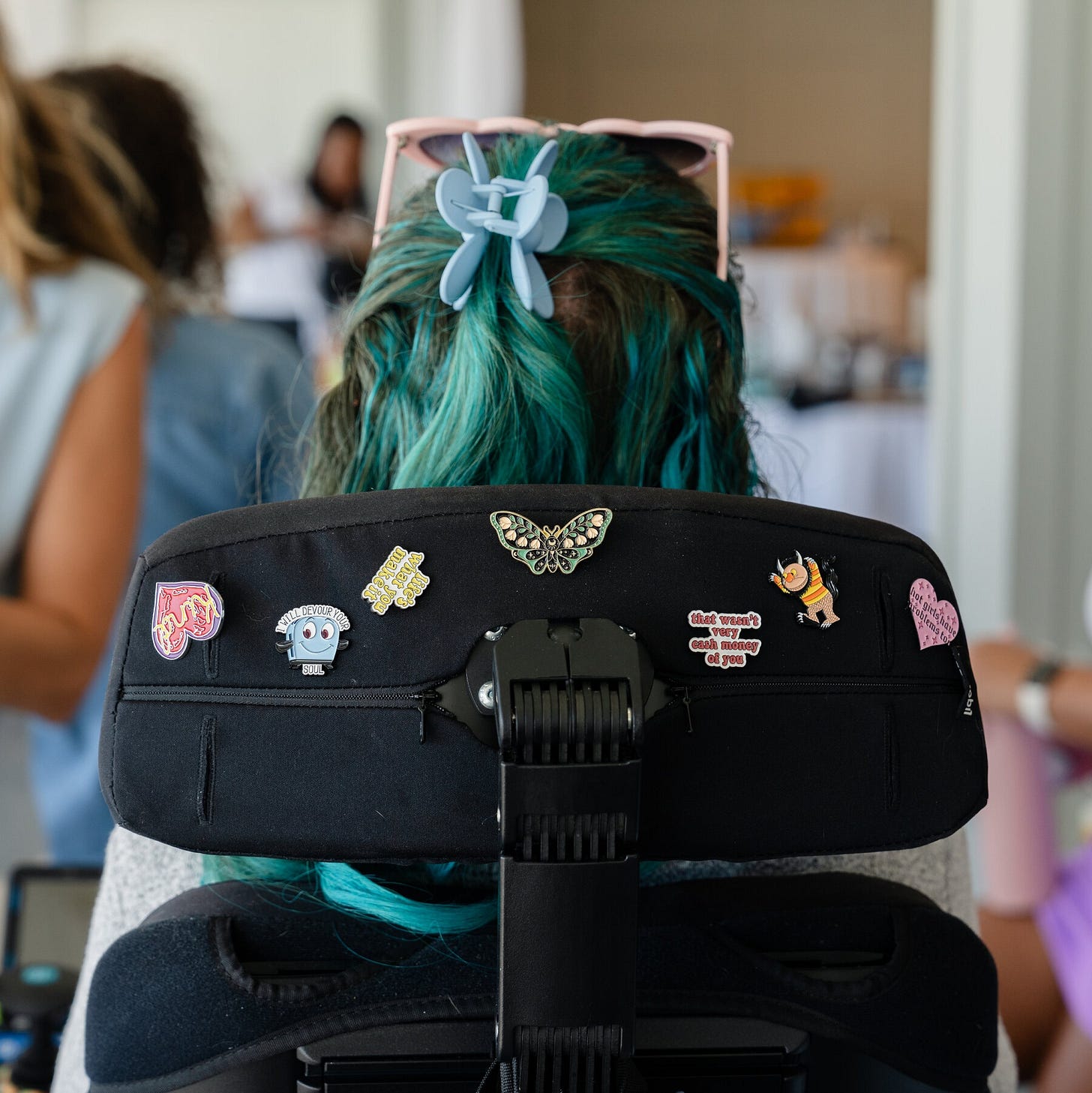 A photo of the back of a woman with teal and brown hair with a blue butterfly clip and pink sunglasses in a powerchair with cute stickers on the back of the headrest.
