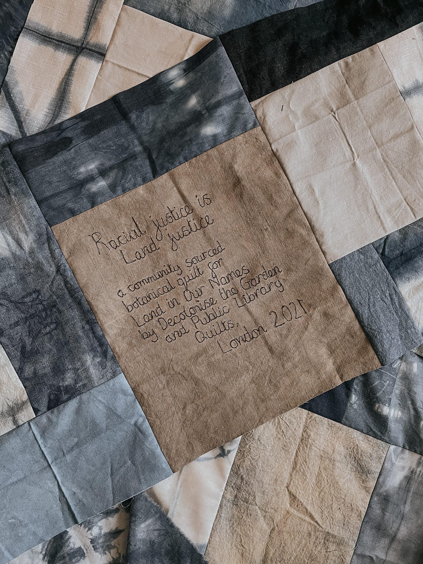 Close up image of quilt with a patch stating: Racial justice is Land justice. A community sourced botanical quilt for Land In Our Names by Decolonise the Garden and Public Library Quilts. London 2021. Close up image of quilt with a patch stating: Racial justice is Land justice. A community sourced botanical quilt for Land In Our Names by Decolonise the Garden and Public Library Quilts. London 2021.