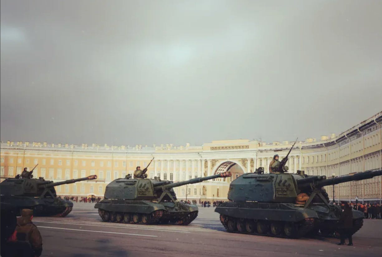 Military tanks with long gun barrels lined up in Palace Square during a parade, positioned in front of the neoclassical columns of the General Staff Building under an overcast grey sky, with soldiers standing on the vehicles.