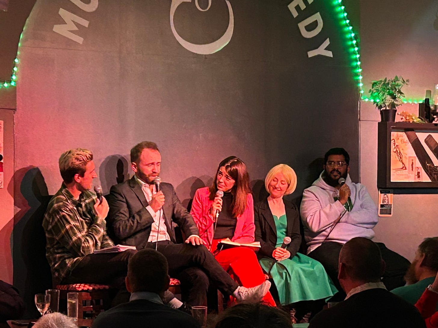 five comedians sit on stools on stage, each holding microphones