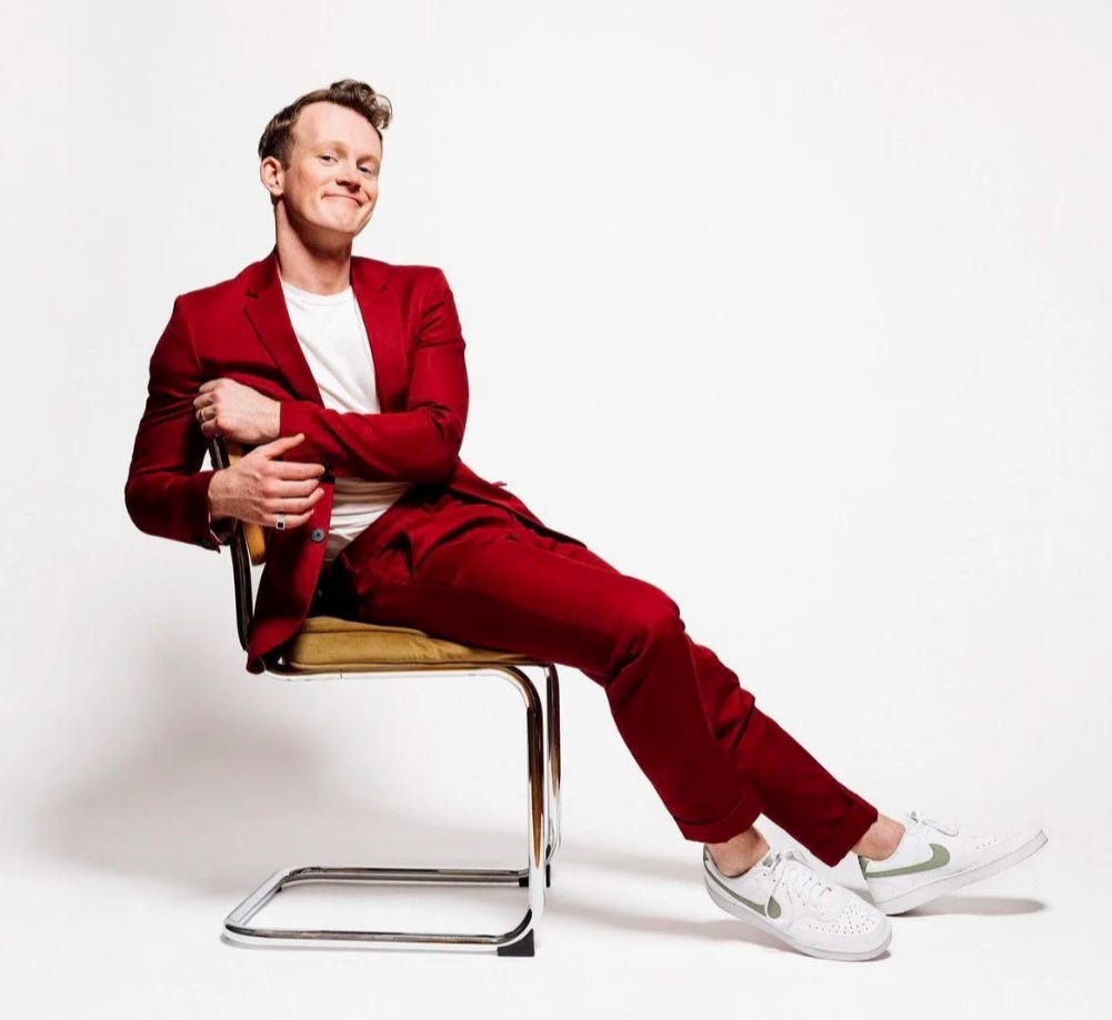 PJ Adzima in a maroon suit, leaning back on a chair in a white studio.