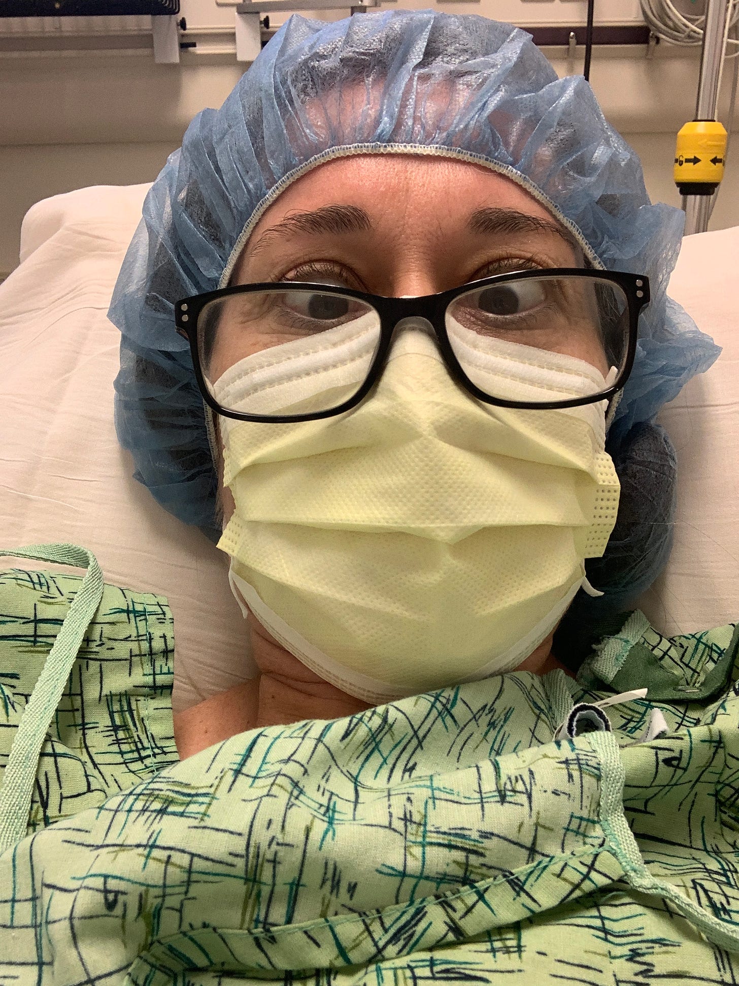 A cross-eyed person laying in a hospital bed wearing a gown, a hair cover, face mask and glasses. 