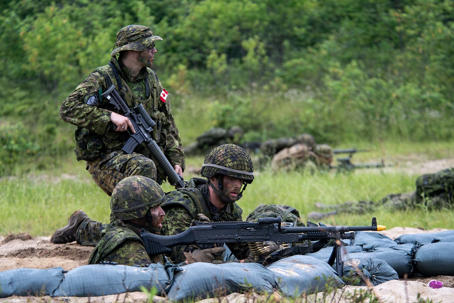 Two members of the Canadian Armed Forces are lying low to the ground, aiming a machine gun at an off-camera target. Another member is kneeling on one knee with their rifle, looking away from the camera.