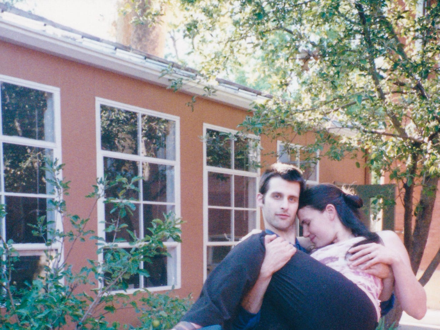 A man carries a woman. He looks at the camera. She has her eyes closed, her forehead to his cheek. Behind them is an orange building with white windows and trees. It’s summer and afternoon.