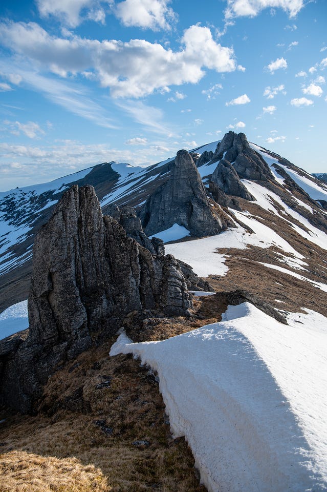 A row of jagged granite outcrops runs along a snowy ridge crest beside a curving snow cornice, under a blue sky streaked with thin cloud.