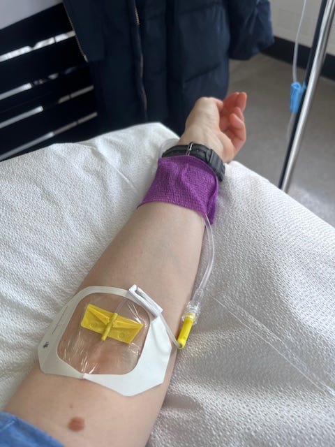 POV shot of OP's extended arm resting on a pillow, palm-up. A clear dressing covers the entrance of the IV line. A band of purple gauze around my wrist wraps around the line to keep it close to my arm, so it doesn't get caught on anything.