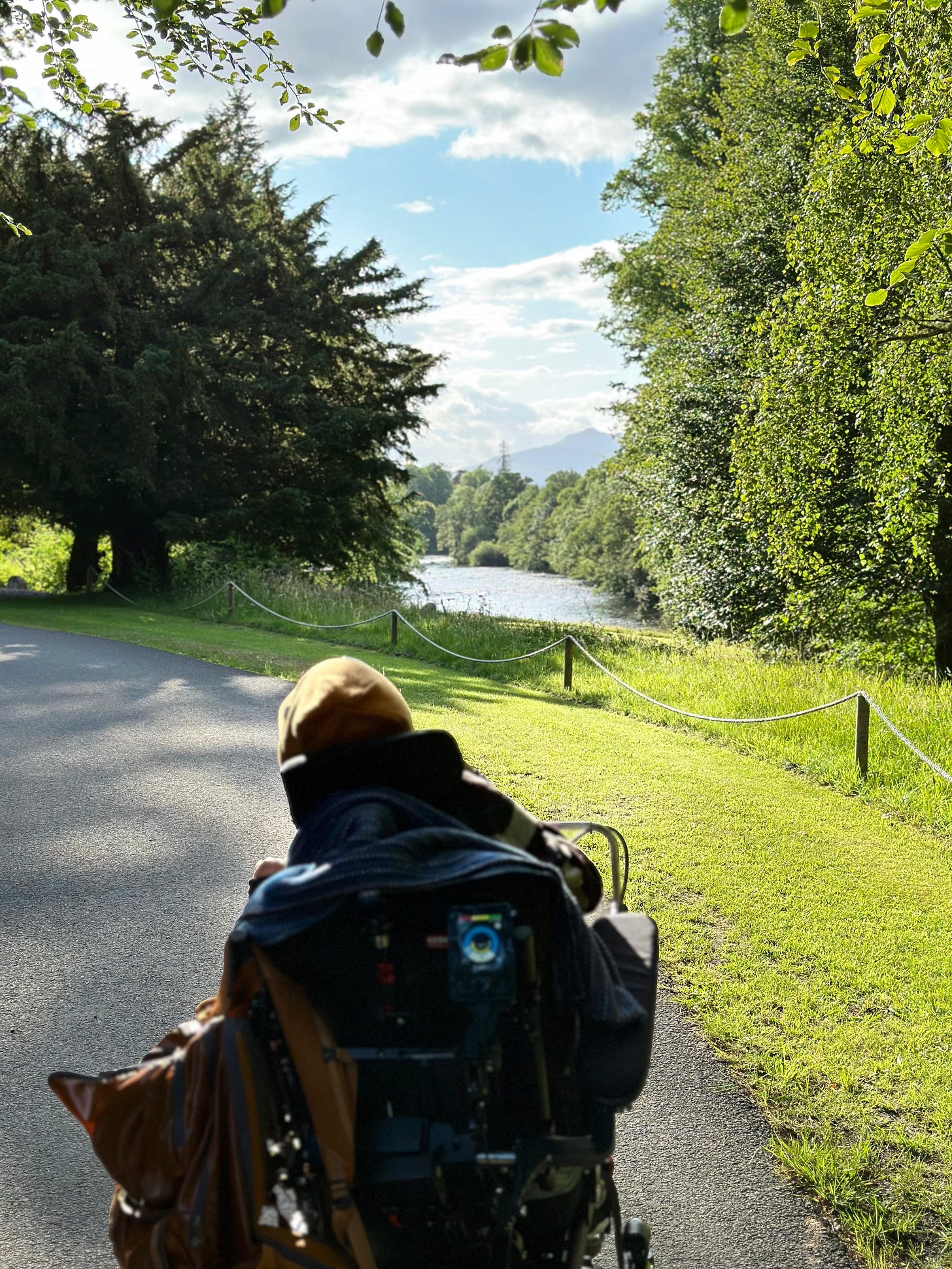Rear view of Sulaiman sitting in his power wheelchair, wheeling along a footpath, exploring nature in the sunshine during our sacred and secret family trip to Scotland between June and July 2023.