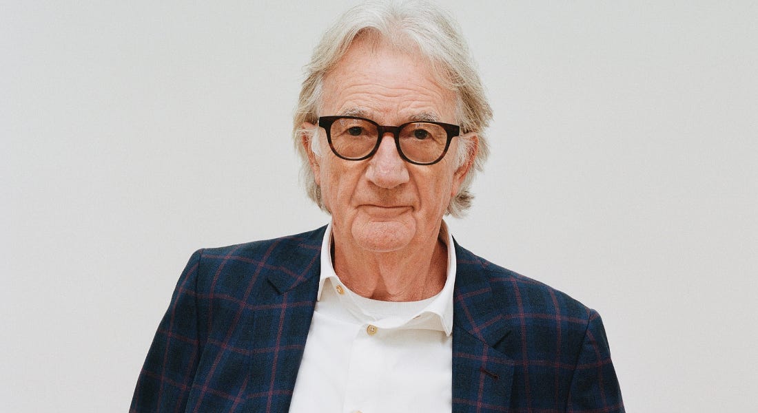 Paul Smith wearing black glasses, a white shirt, and a blue and purple blazer, standing in front of a grey background Paul Smith wearing black glasses, a white shirt, and a blue and purple blazer, standing in front of a grey background