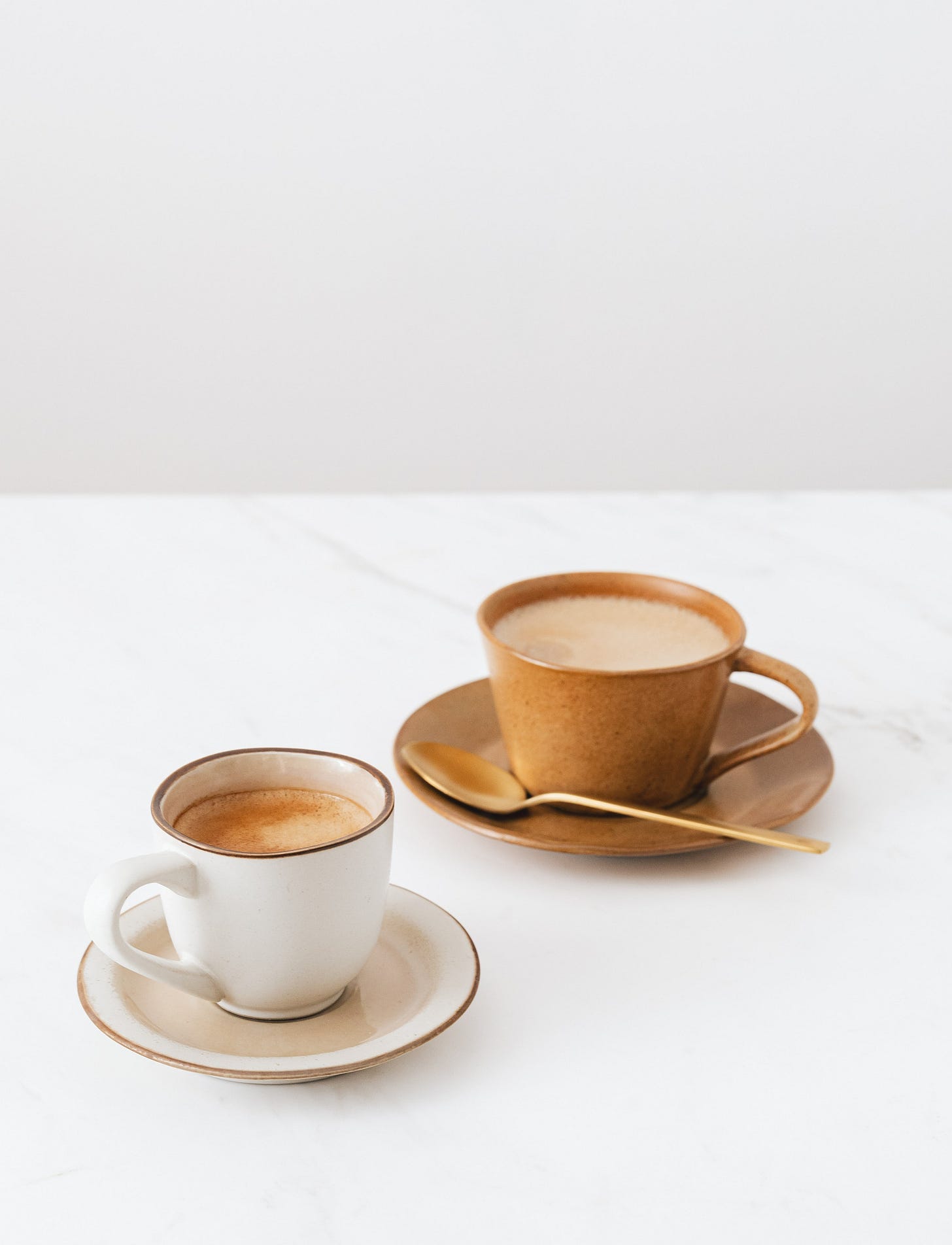 two light brown cups of coffee on saucers with golden spoons on white table and white background