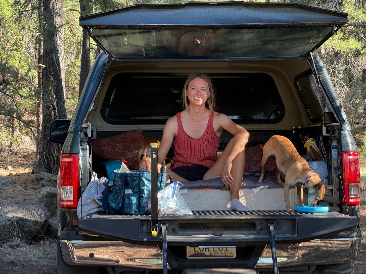 a woman sitting in a truck bed set up with a mattress, pillows, bags for cooking supplies, with her dog next to her eating on the tailgate