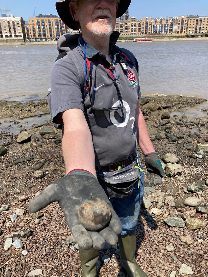 Man holding a canon ball, a shark's tooth and a typical mix of stones and pebbles on the banks of the river Thames