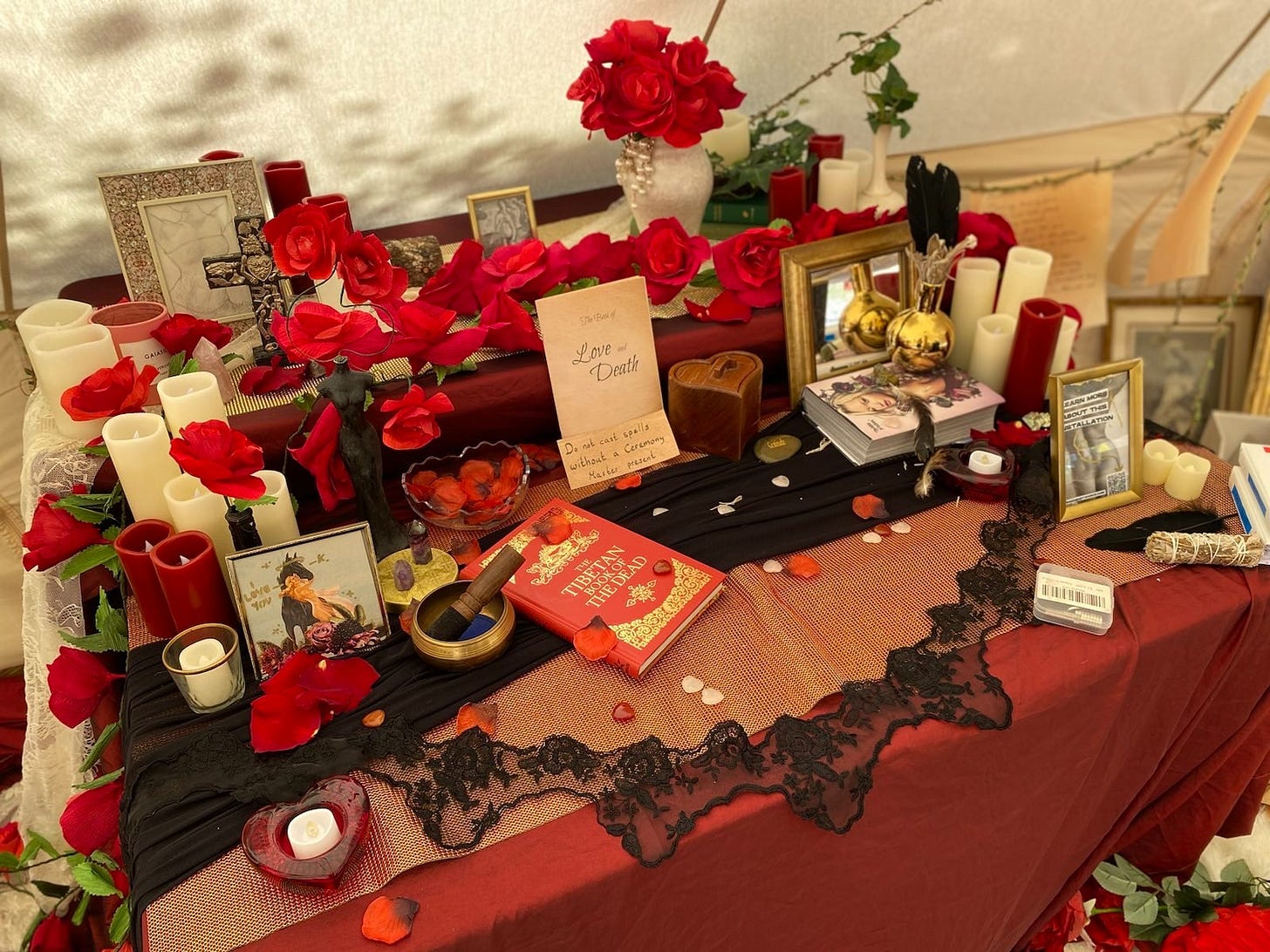 A photo of an altar table covered in red roses and (LED) candles, small ornamentation, photo frames, and a book titled "The Book of Love and Death", and a book The Tibetan Book of the Dead
