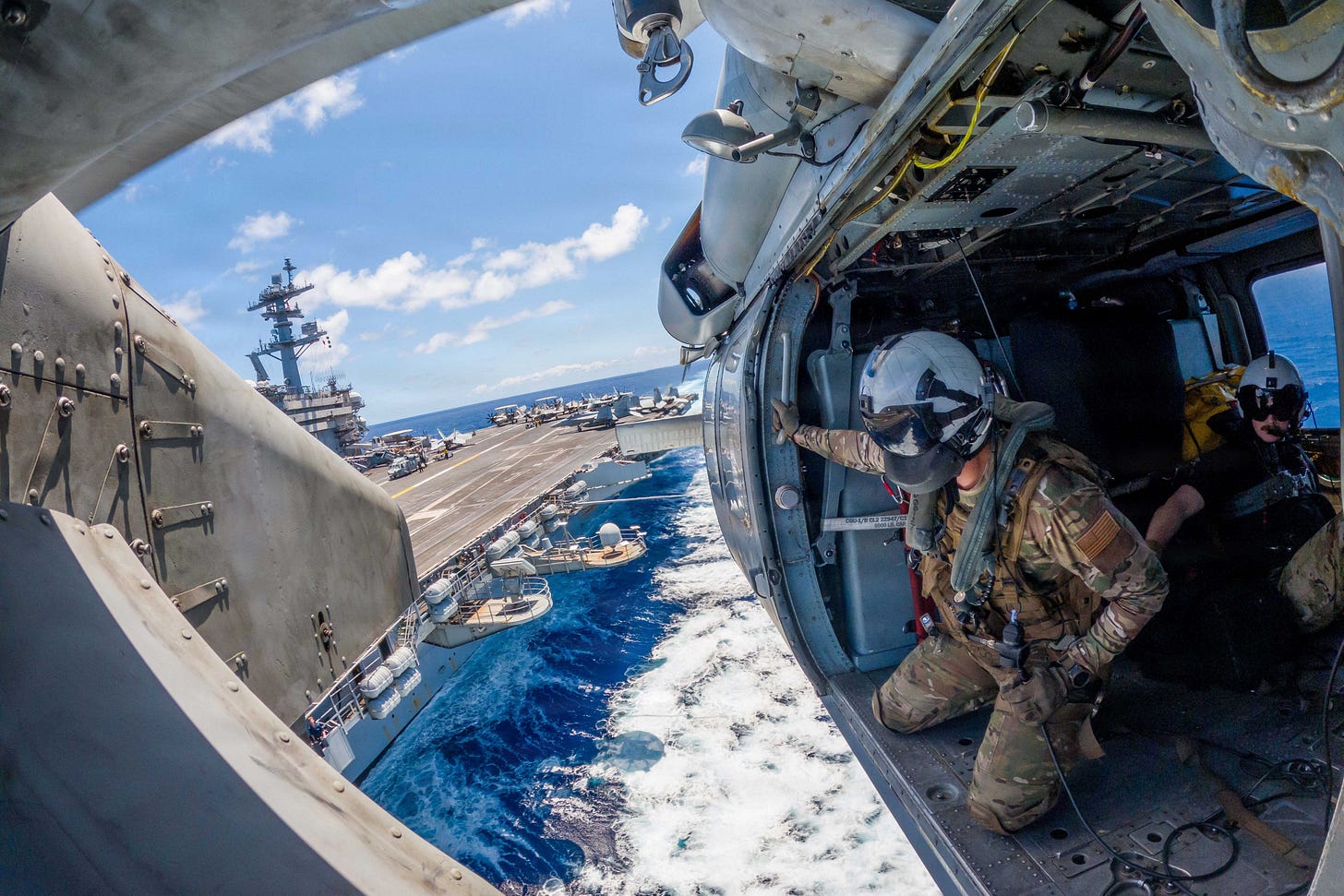 Navy aircrew looking out an open helicopter door toward an aircraft carrier and its jets at sea Navy aircrew looking out an open helicopter door toward an aircraft carrier and its jets at sea