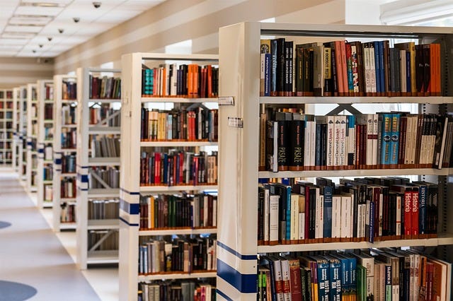 library shelves with stacks of books, via Pixabay, in the public domain
