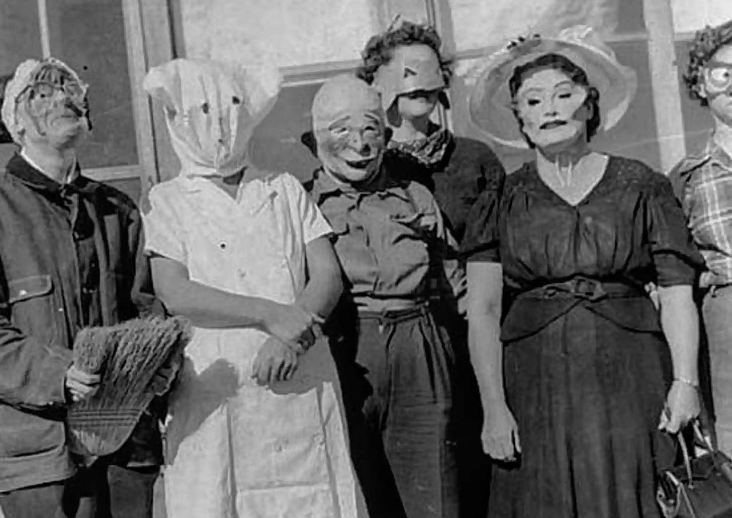 A vintage black and white image of women dressed in handmade halloween costumes. They wear nurse's dresses and men's worksuits, and a black dress, some have cheap rubber masks, others have paper cut out masks and one has a bag with holes for the eyes and mouth