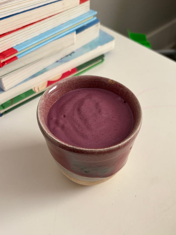 A toddler hand reaching for an unblended smoothie poised beneath a blue and green miniature excavator. A blended smoothie in a ceramic cup next to a stack of books.