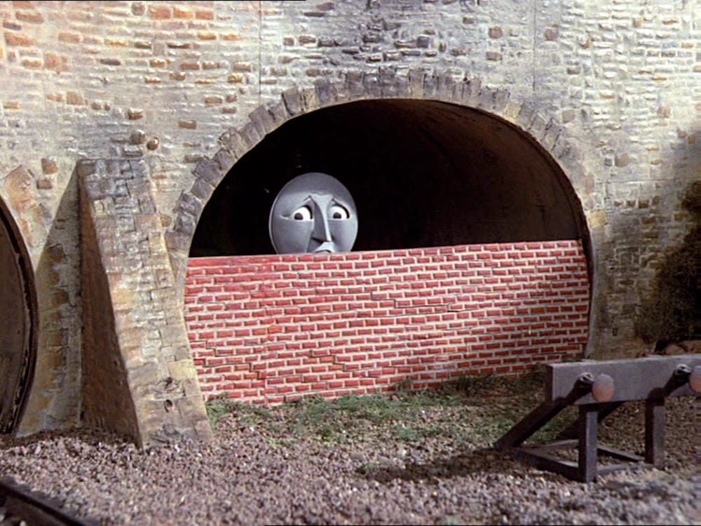 A model anthropomorphic train with a gray face and sad eyes peers out from a tunnel as it is bricked up with him inside. The tunnel interior is very dark. The exterior is bright and sunny and the plastic of the model tunnel shines slightly.