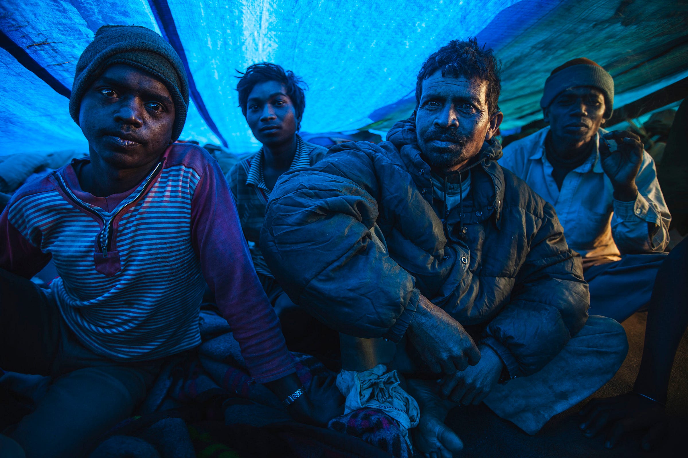 Four men sit closely together under a blue plastic tarp, illuminated by blue light. They wear warm clothing and have serious expressions. The setting appears makeshift, suggesting shelter or temporary accommodation. Four men sit closely together under a blue plastic tarp, illuminated by blue light. They wear warm clothing and have serious expressions. The setting appears makeshift, suggesting shelter or temporary accommodation.