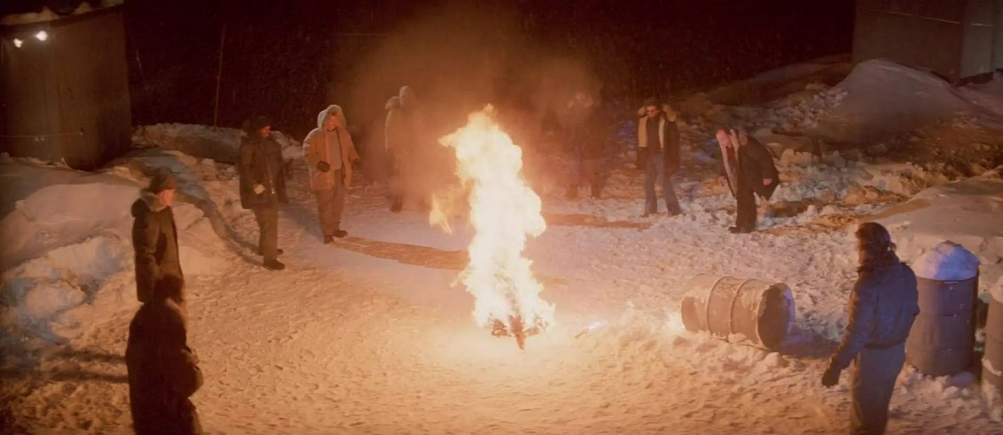 A group of men are outside in the snow, standing around a fire in the middle of the circle.