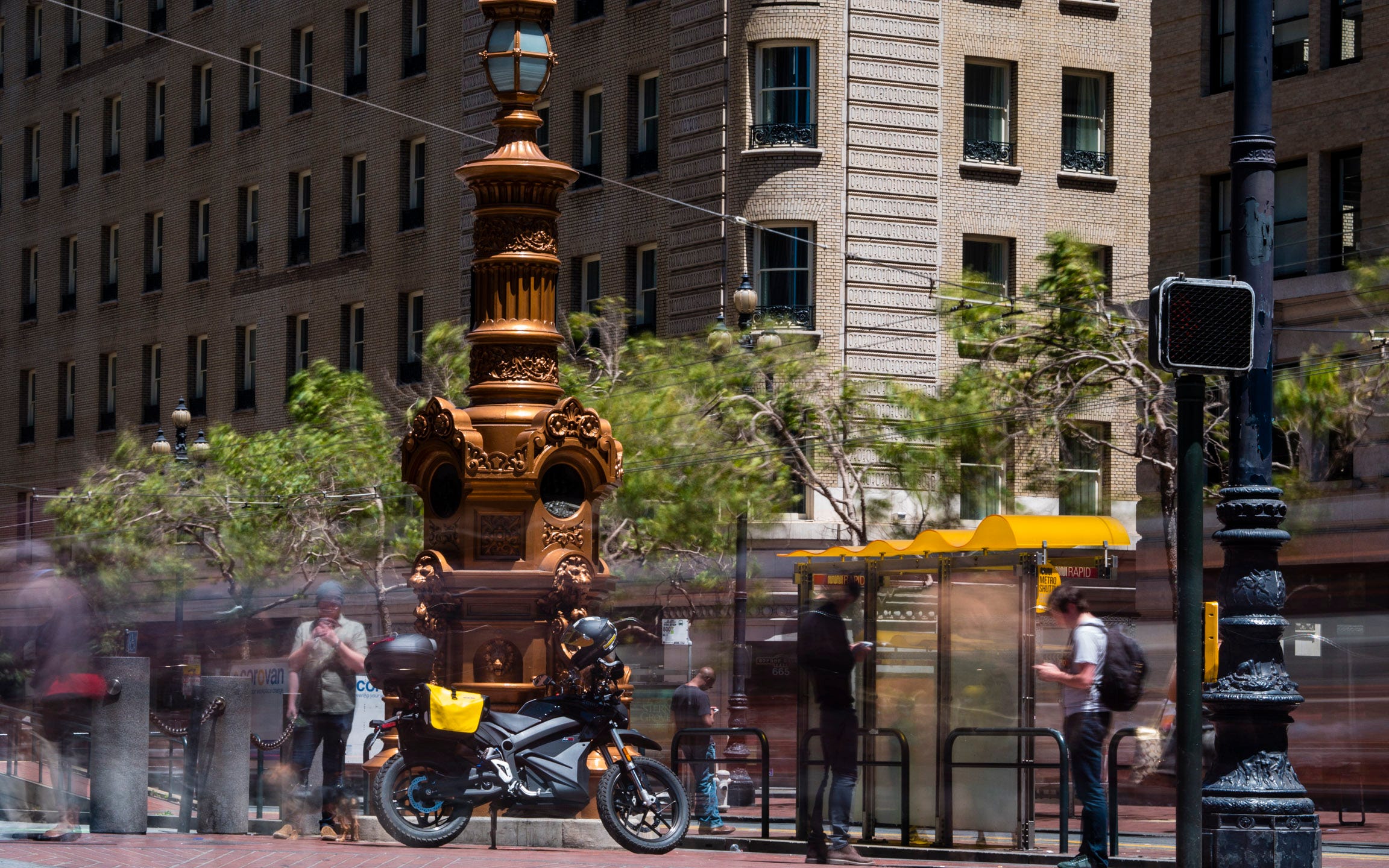 Closeup photo of Lotta's Fountain, an ornate, early 20th century bronze statue. It is approximately 20 feet tall and is cylindrical in form. The first ten feet is eight-sided, with small cast lion's heads, arched niches, and small basins where water once flowed. Above, the statue tapers into a stout, fluted column with a faceted lamp at its peak. In front of the statue sits the Zero DSR electric motorcycle with yellow saddlebags and a black topcase. People stand around the fountain and bike looking at their phones. Those that are moving during the long exposure of the photo appear as blurs. 