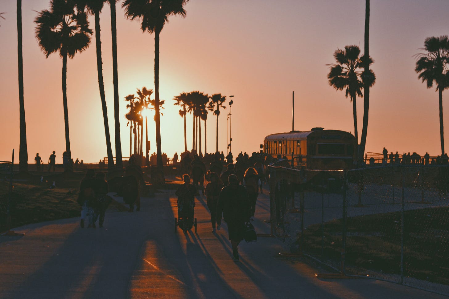 People walk along Venice Beach at sunset, silhouetted against a vibrant sky with palm trees and a bus. People walk along Venice Beach at sunset, silhouetted against a vibrant sky with palm trees and a bus.