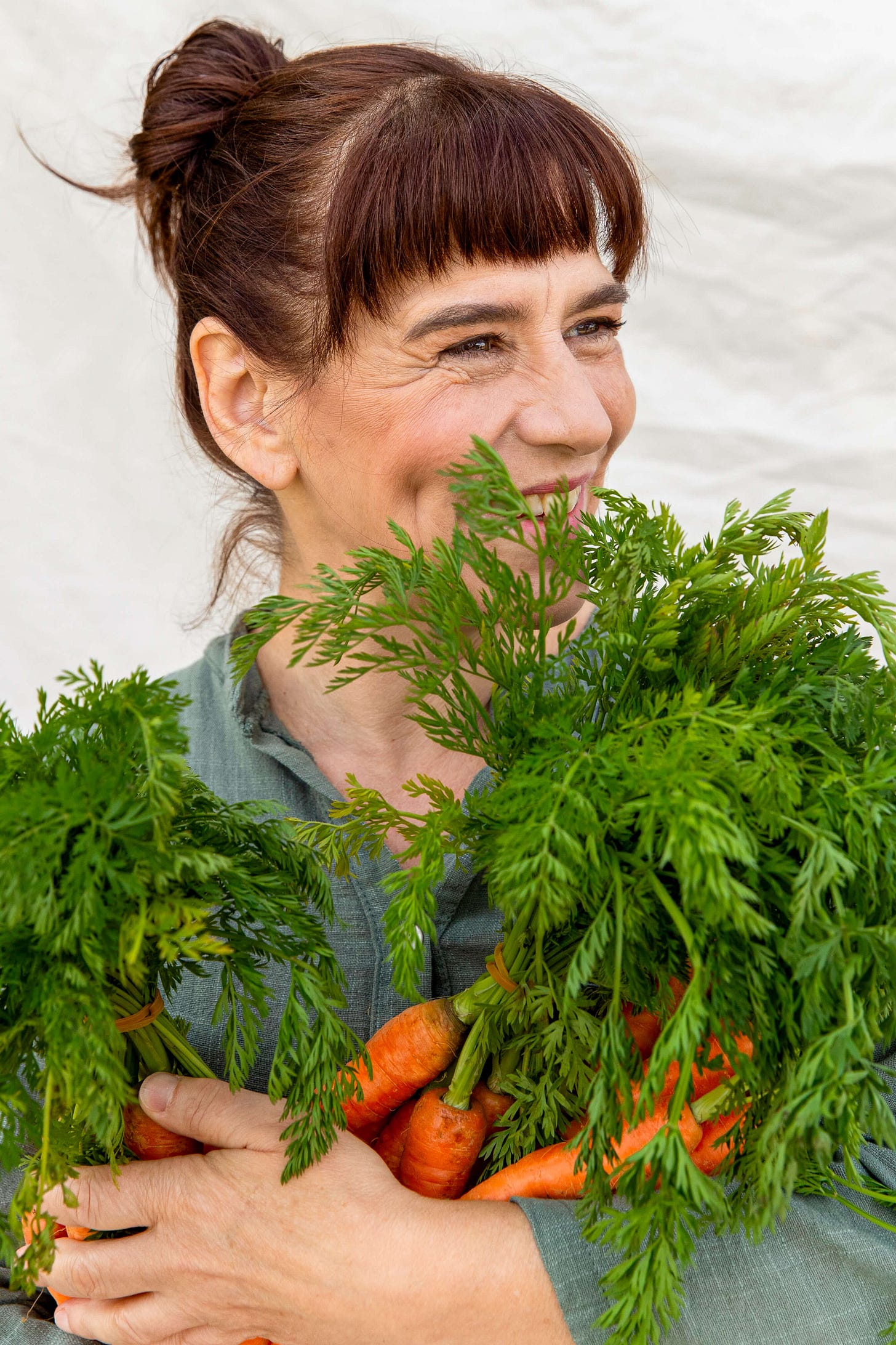 Die Autorin Esther Kern lächelt und hält mehrere grosse Bündel frischer Karotten mitsamt üppigem Grün im Arm. Sie trägt ein grünes Shirt, steht vor einem hellen Hintergrund und wirkt, als würde sie den Duft des Karottenkrauts geniessen.