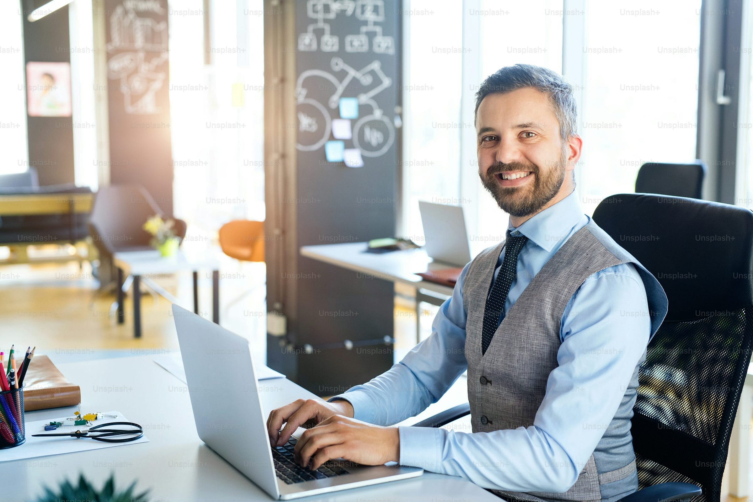 Handsome young businessman in his office, sitting at the desk, laptop in front of him, smiling Handsome young businessman in his office, sitting at the desk, laptop in front of him, smiling
