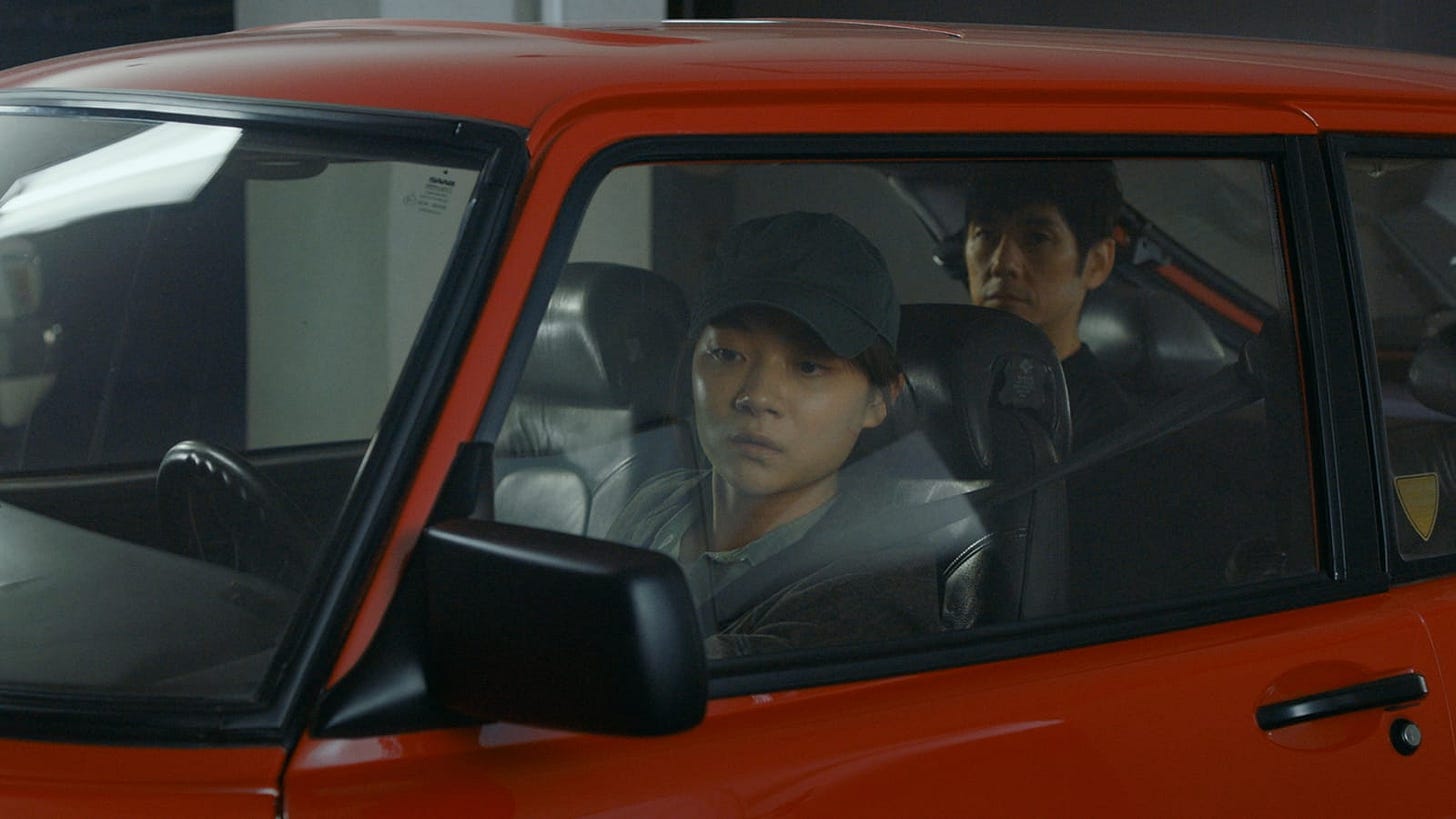 Drive My Car | Still features Hidetoshi Nishijima as Yusuke Kafuku as a passenger in a red car being driven by Toko Miura as Misaki Watari.