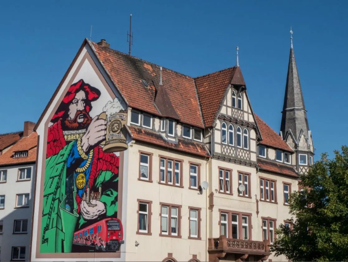 StreetArt mural by YoungArt on a house in Einbeck, Germany. The artwork shows a bearded man in red holding a large beer stein, with bold colors and modern design elements. The mural honors Einbeck’s famous Bockbier brewing tradition.