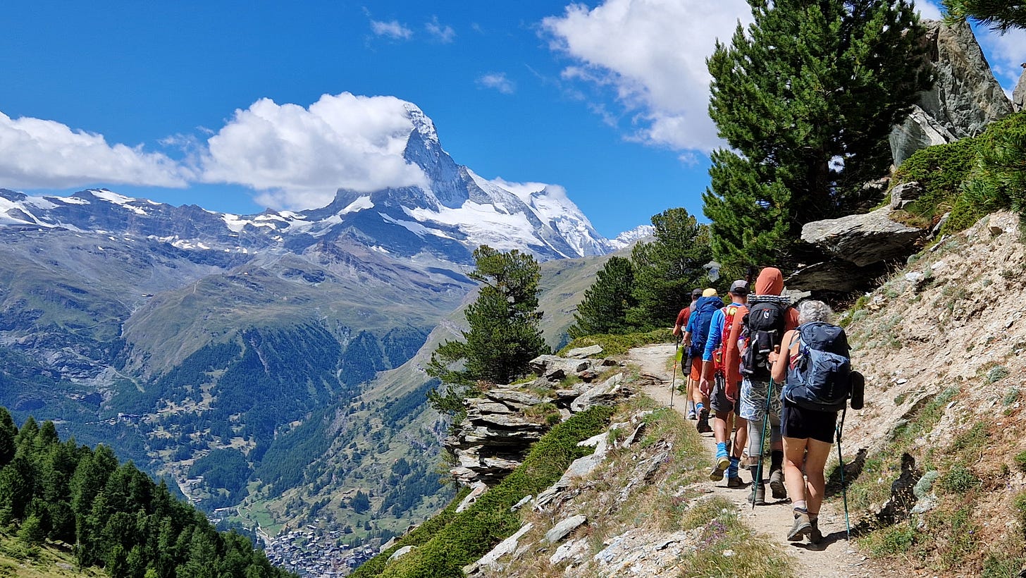 Hikers walking along a narrow path with The Matterhorn in background partially covered in clouds. Blue sky, sunny Hikers walking along a narrow path with The Matterhorn in background partially covered in clouds. Blue sky, sunny