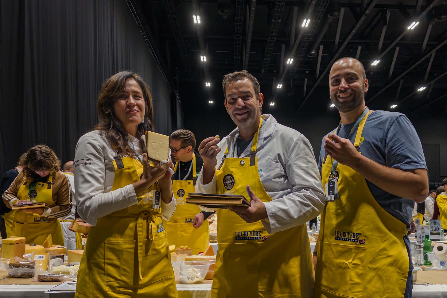 Drei Jury-Mitglieder der World Cheese Awards 2025 stehen in einer grossen Halle vor einem Tisch voller Käseproben. Alle tragen gelbe Schürzen und halten Stücke Käse in der Hand; die Frau links präsentiert einen halben Laib, die beiden Männer daneben probieren jeweils ein kleines Stück. Im Hintergrund arbeiten weitere Personen an ihren Bewertungstischen.