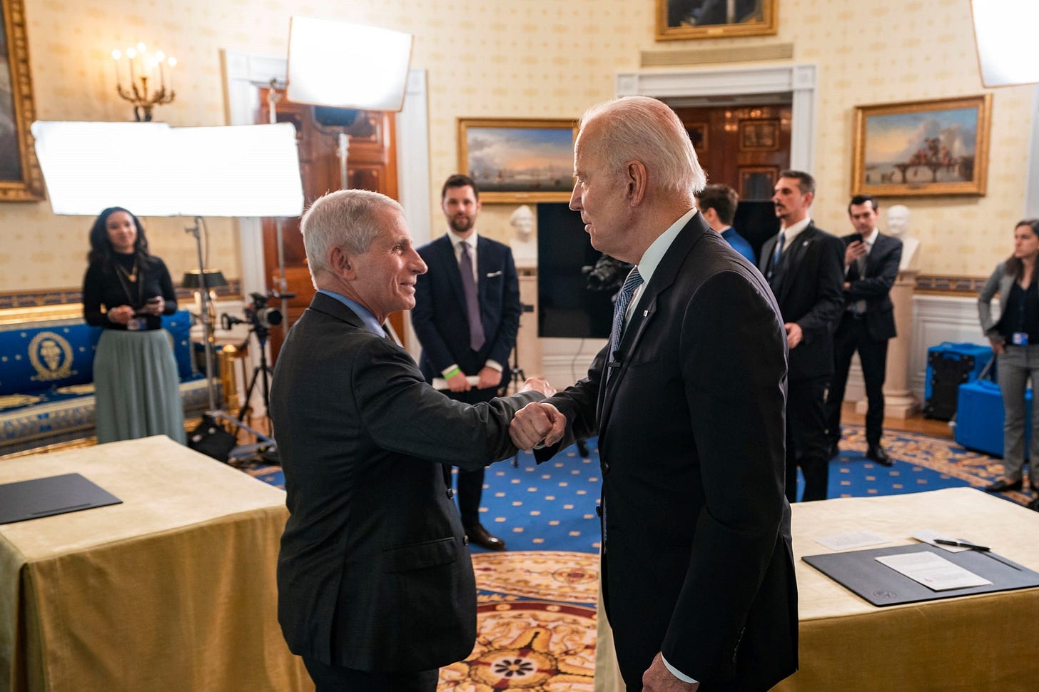 Dr. Anthony Fauci shakes hands with President Joe Biden in a formal White House room with staff and equipment behind them.