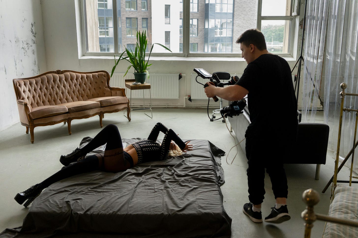 A photography session in progress within a bright studio with large windows. A photographer operates camera equipment while photographing a model posing on a dark gray floor mat. The space features a vintage beige tufted sofa, white walls, and natural lighting from large windows.