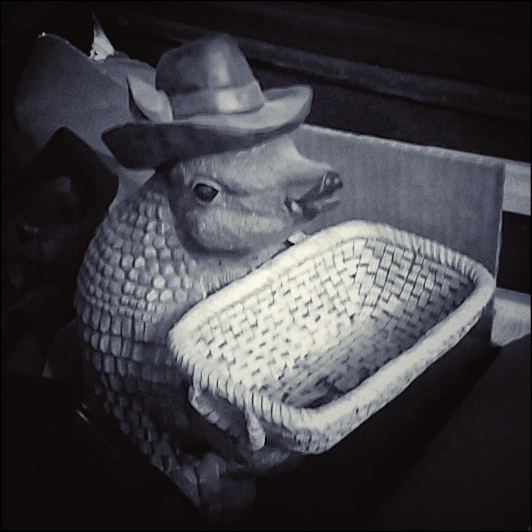 A monochrome photograph of what appears to be an armadillo sitting on its haunches. The armadillo is wearing a hat -- a fedora, with its ears poking up through the brim -- and holding in its forepaws a woven basket. There's something in the bottom of the basket, but it's too small to identify.