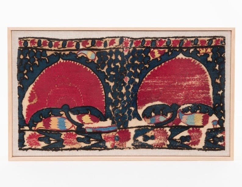 May include: Framed textile art featuring two large red arch shapes against a dark blue background with a floral design. The artwork has a light-colored wooden frame and a beige fabric border. The design includes accents of yellow, blue, and red.