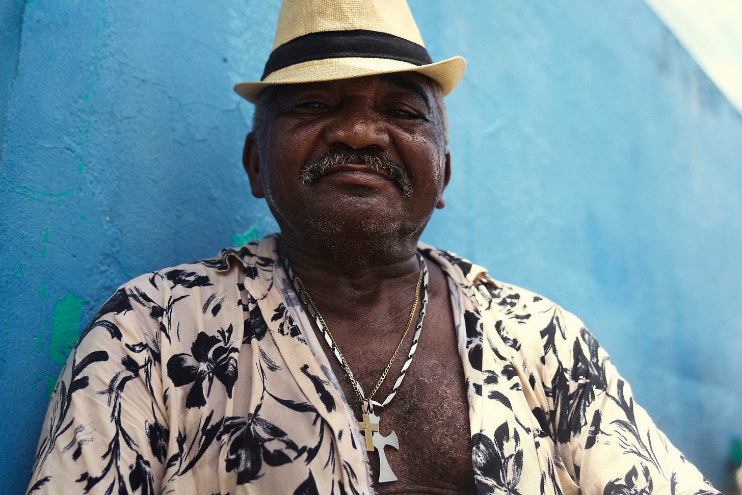 An older man wearing a straw hat, floral shirt, and cross necklace sits against a textured blue wall, looking at the camera with a slight smile.