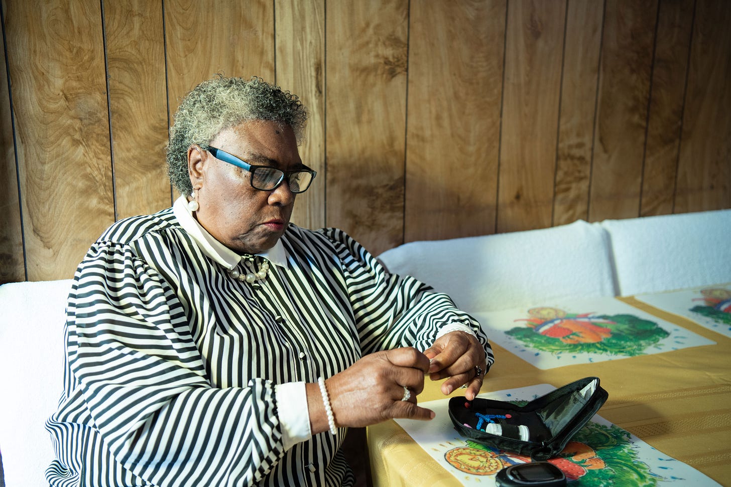 An image of a woman sitting at a table preparing to test her blood glucose. She has short curtly hair and glasses, and wears a black and white striped blouse. A zippered pouch sits on the table beside her. An image of a woman sitting at a table preparing to test her blood glucose. She has short curtly hair and glasses, and wears a black and white striped blouse. A zippered pouch sits on the table beside her.
