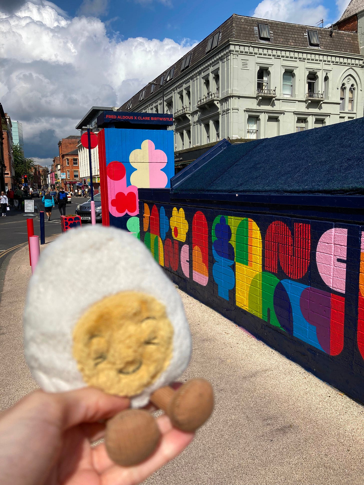 Photograph of dippy the soft toy egg in front of a sunny street in Manchester, covered in street art of abstract shapes in pretty colours. 