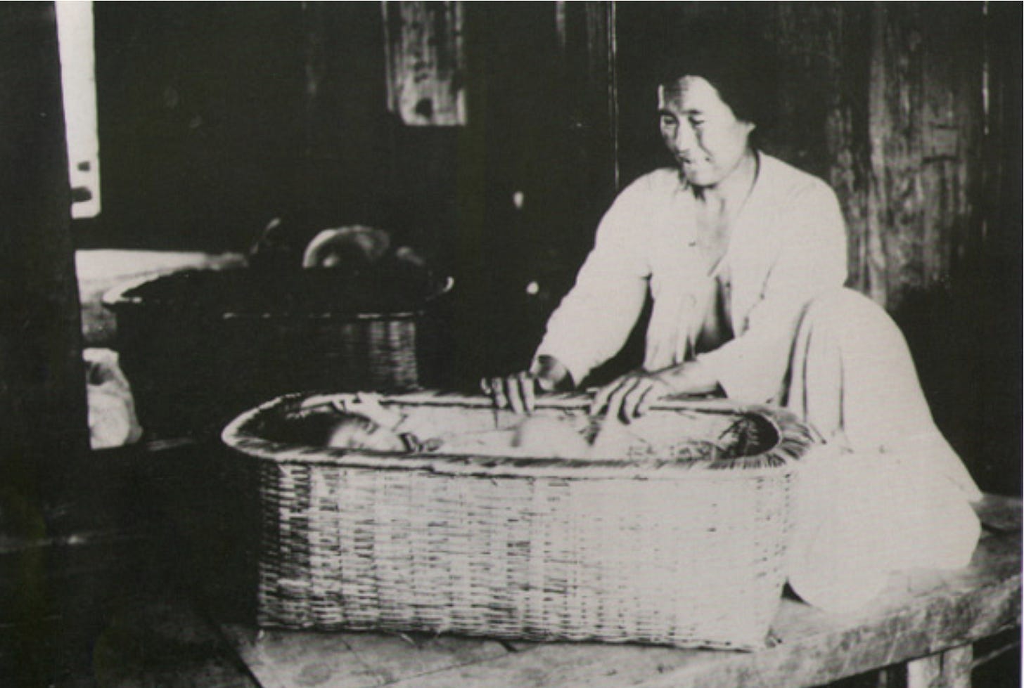 A vintage, black and white photograph shows an older Korean woman, dressed in traditional attire, sitting cross-legged on a raised wooden platform. She is smiling down and has her hands on the side of a rectangular, intricately woven bamboo basket (aegi gudeok) that serves as a cradle for a baby sleeping inside it. The woman sits in a traditionally constructed wooden-frame room, with dark wood beams and planks visible. In the background, out of focus, are two larger, deep woven baskets. The scene is quiet, intimate, and evokes a sense of historical daily life.