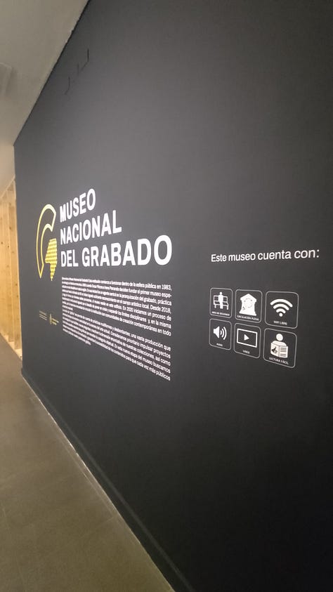 1- Fotografía de una cartelería externa de un museo, hecha de estructura de metal de aproximadamente unos tres metros de altura y sesenta centimetros de ancho. Se encuentra ubicado en el medio de un parque, en un sector donde hay tránsito peatonal. En la cartelería hay pictogramas que indican los espacios, servicios, contenidos y actividades de el museo castagnino 2- Fotografía de una cartel externa de un museo ubicado en el jardin previo al ingreso al edificio. El cartel es un rectángulo horizontal con un pie metalico que entra en el suelo. Da la bienvenida y expone pictogramas que indican los espacios, servicios, contenidos y actividades con los que cuenta el museo. 3- Fotografia de una pared en el interior de un museo. la pared esta pintada de negro y tiene en color blanco la leyenda con el nombre dle museo nacional del grabado y un texto. Al costado derecho hay un listado de pictogramas que indican los espacios, servicios, contenidos y actividades con los que cuenta el museo. 4- Fotografia de una pared en el interior de un museo. la pared es blanca y tiene en el centro un recuadro con borde negro y en su interior un texto que da la bienvenida. Debajo hay un listado de pictogramas que indican los espacios, servicios, contenidos y actividades con los que cuenta el museo. 5- Fotografia de un sector de una exhibición. En la pared hay cuatro imagenes enmarcadas y debajo en un exhibidor hay folios aillados con documentos antiguos y otras imagenes. En un costado se encuentra un pictograma y un código qr. 6-Fotografia de una sala de exhibición de un museo de arte contemporaneo. En el centro hay una mujer de pie agarrando el celular con las dos manos para sacar una fotografia a un dispositivo que hay en una de las paredes de la exhibición. detrás de ella, al final de la sala hay mas personas de pie y sentadas.