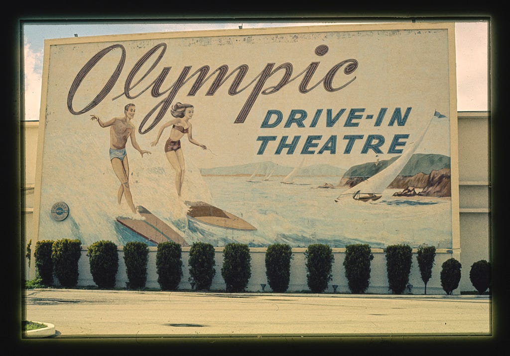 Large billboard with text "Olympic Drive-In Theatre" on the side a building with a green hedge in front. Large billboard with text "Olympic Drive-In Theatre" on the side a building with a green hedge in front.