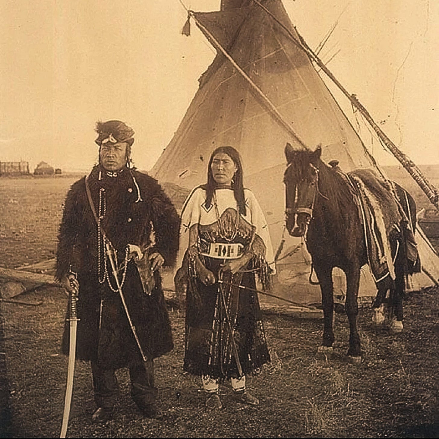 Photo of Blackfoot warrior standing next to wife and horse, in front of teepee, and holding a katana sword.