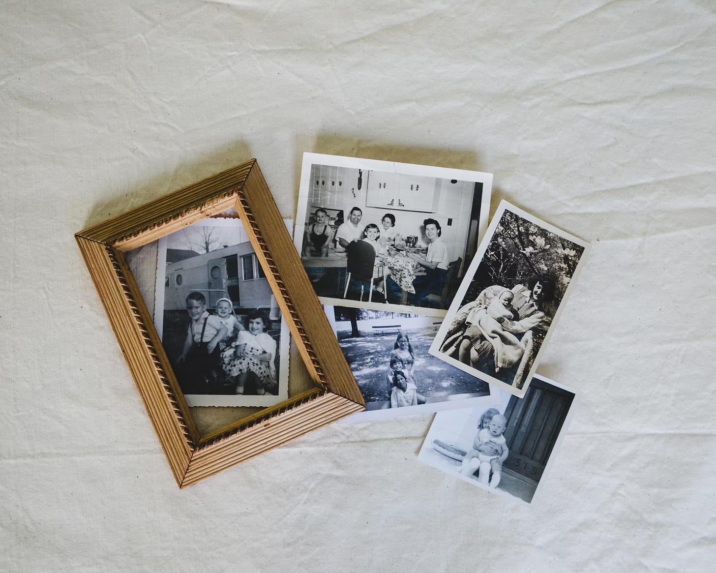 A collection of artistically arranged old family pictures, mostly of children (some featuring adults, presumably parents), arranged on a cream-colored sheet and partially covered by a wood picture frame. A collection of artistically arranged old family pictures, mostly of children (some featuring adults, presumably parents), arranged on a cream-colored sheet and partially covered by a wood picture frame.