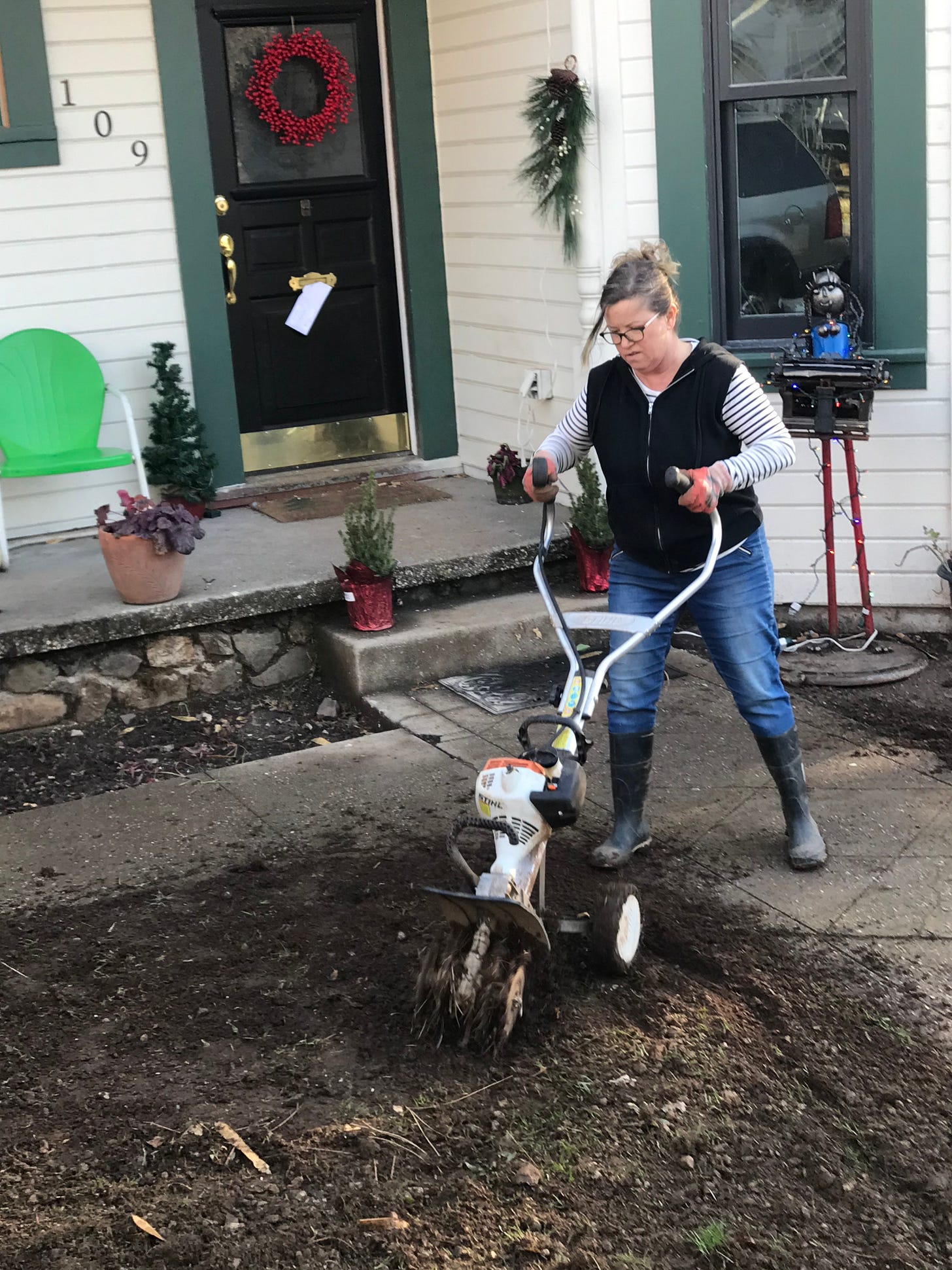 White woman in rubber boots and jeans rototilling patch of dirt in front of a white and green house.