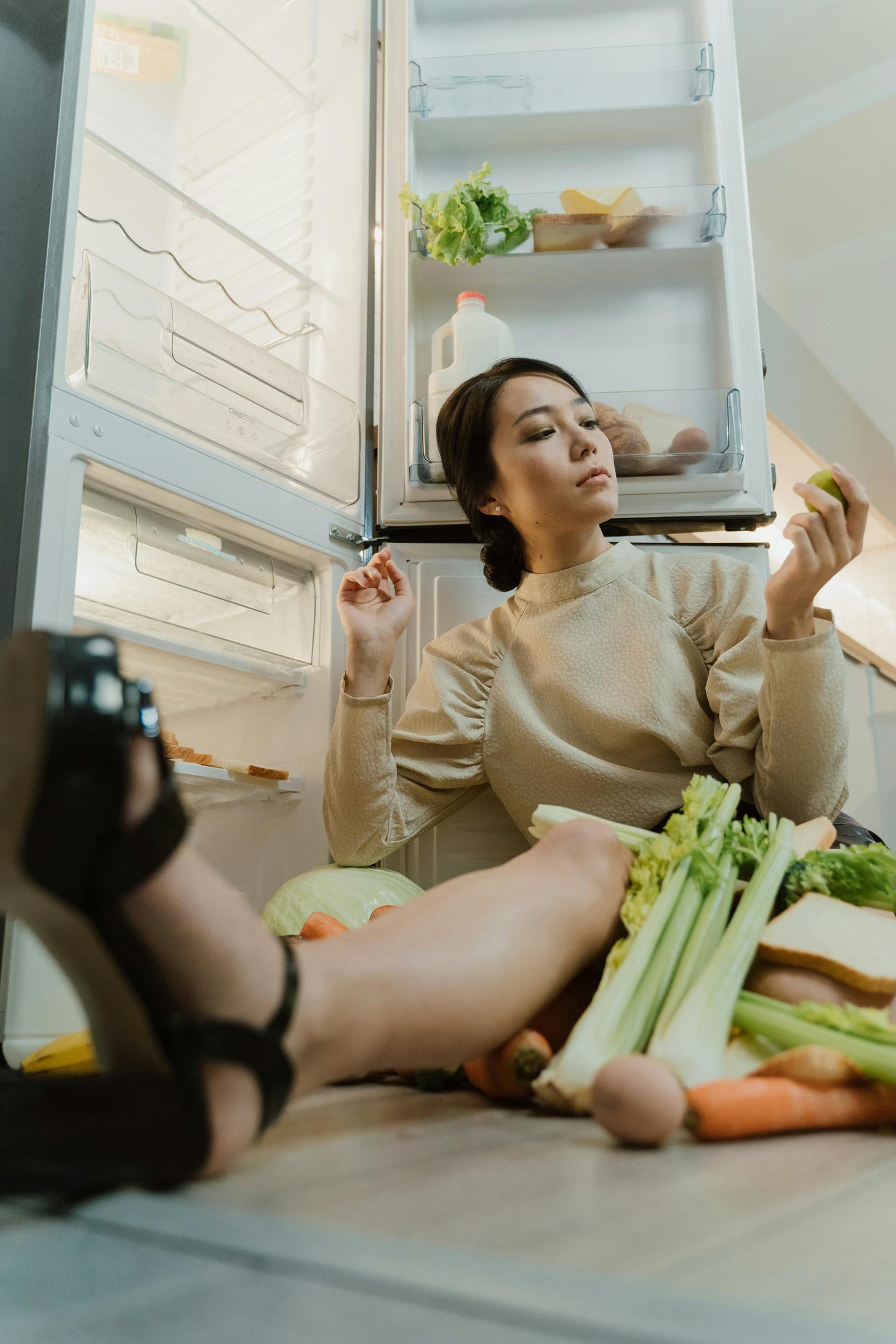 A woman sitting on the floor in front of an open refrigerator, surrounded by various fresh vegetables and other groceries, holding a piece of produce and looking at it. A woman sitting on the floor in front of an open refrigerator, surrounded by various fresh vegetables and other groceries, holding a piece of produce and looking at it.
