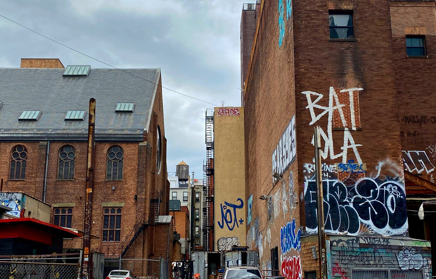 The brick power station on the right. On the left, on the other side of what looks like a parking lot with a dumpster is the back of an brick building with orante windows. Between them further back is a water tower. The brick power station on the right. On the left, on the other side of what looks like a parking lot with a dumpster is the back of an brick building with orante windows. Between them further back is a water tower.