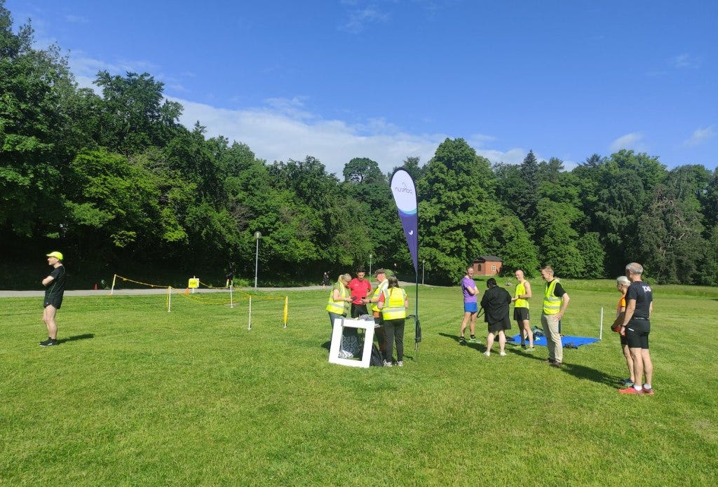 A parkrun flag flying on the grassy area that makes up the finish line. Trees line the path behind. A few runners and several hi-viz volunteers are gathered.