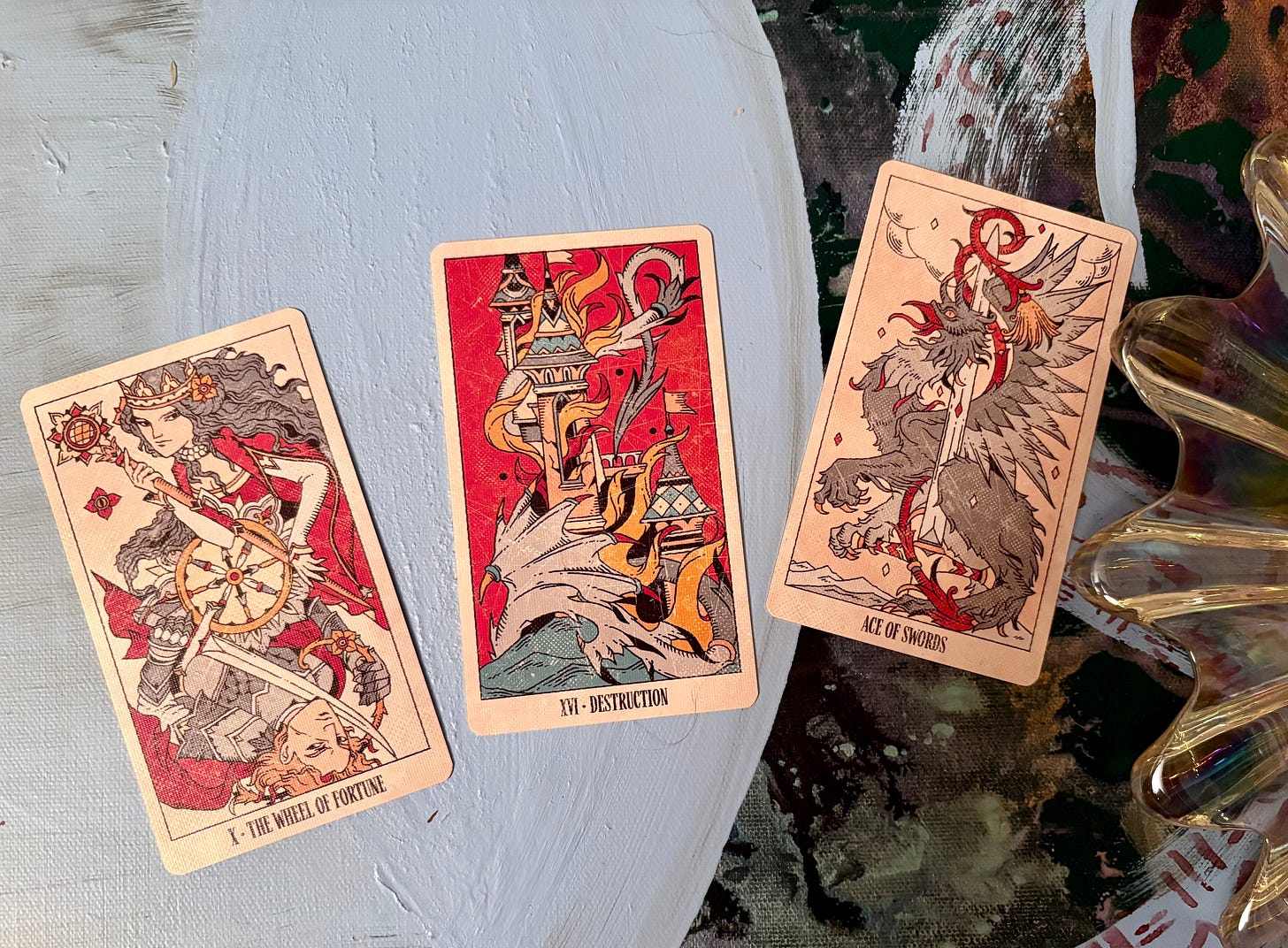 Three tarot cards on a piece of abstract art with a glass bowl to one side. The cards are left to right, X Wheel of Fortune, XVI Destruction, and Ace of Swords Three tarot cards on a piece of abstract art with a glass bowl to one side. The cards are left to right, X Wheel of Fortune, XVI Destruction, and Ace of Swords