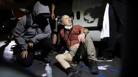 Associated Press A man wearing a t-shirt, trousers and trainers sits on the pavement, one trouser leg rolled up showing gauze wrapped around his right thigh. He wears a camera around his neck and a respirator mask pulled down. Two people look on to him. 
