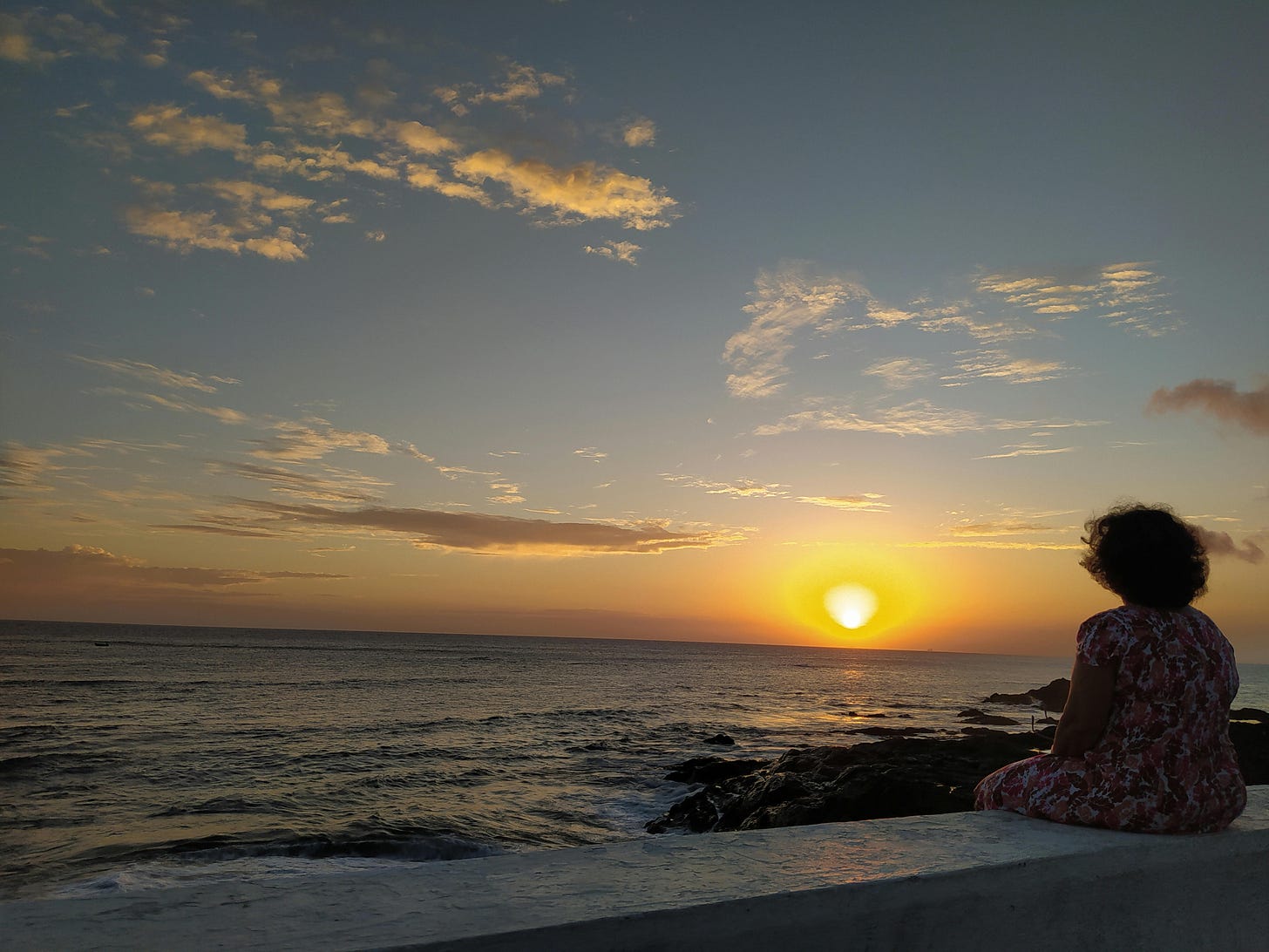 Mulher sentada em um pequeno muro de frente para o mar com o por do sol, a penumbra da tarde se encontra com a noite e as cores se entrelaçam com o bramido das aguas e as núvens amarelas do ceu.